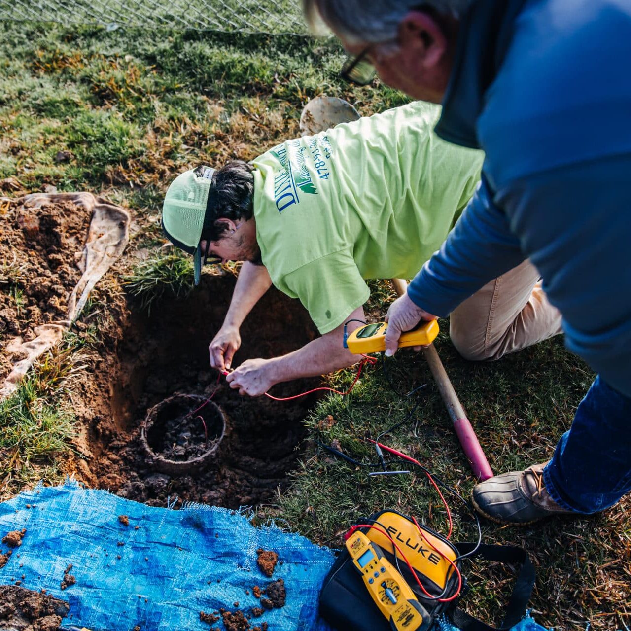 Two people work near a dug hole. One is crouched, examining cables with a tool, while the other stands holding equipment. A blue tarp, tools, and a measuring device are nearby. They're wearing casual outdoor clothing.