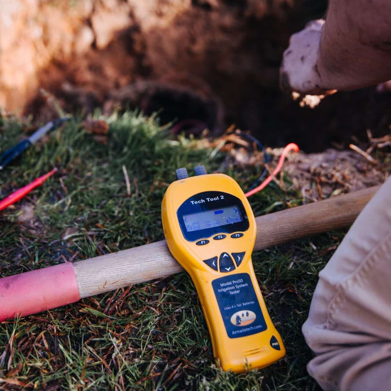 A yellow handheld device labeled Tech Tool 2 rests on the ground near a hole, with red and white wires nearby. A person, partially visible, works beside it on grass.